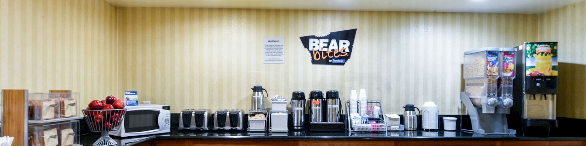 The image shows a breakfast station in a hotel with coffee dispensers, a juice machine, a microwave, a toaster, a cereal dispenser, and a 