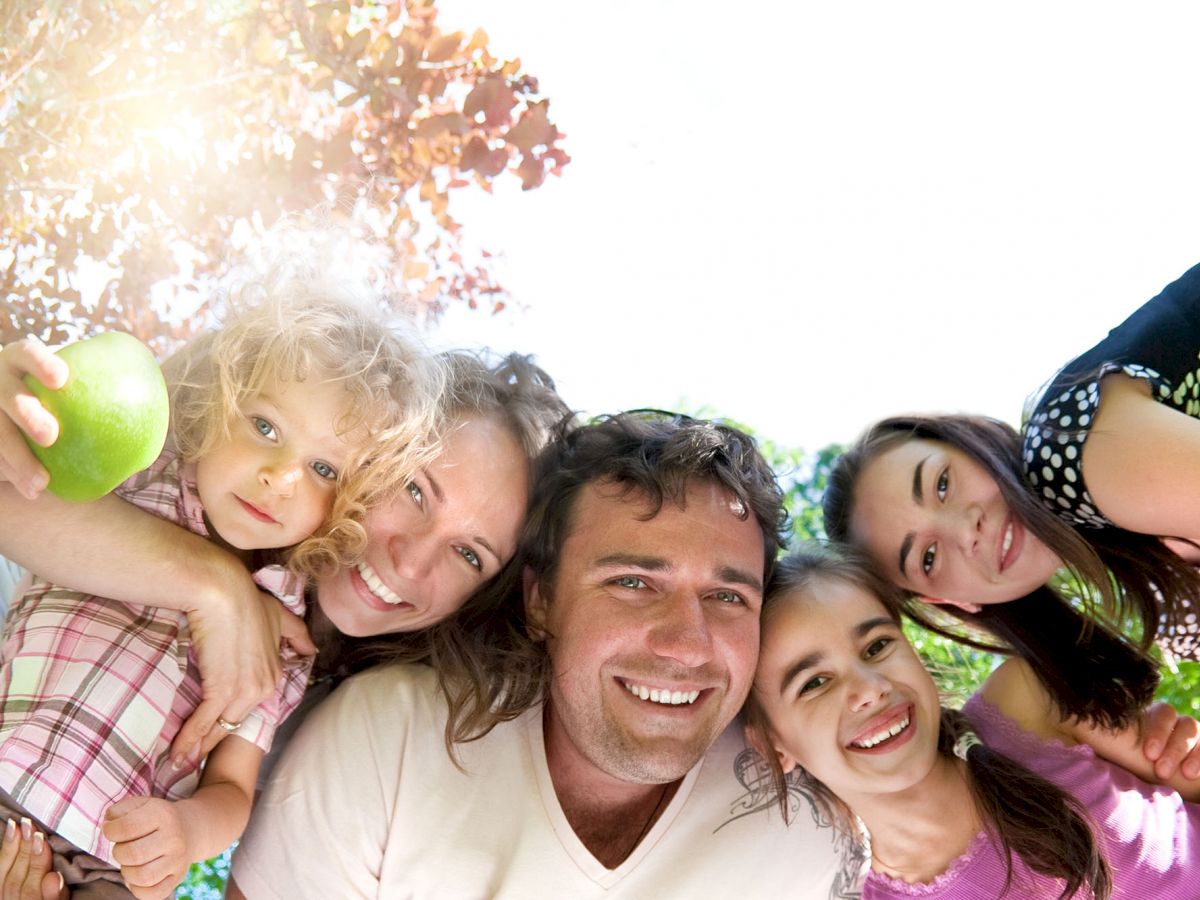 A group of five people posing happily outdoors, with greenery and sunlight in the background, one child holding a green apple.