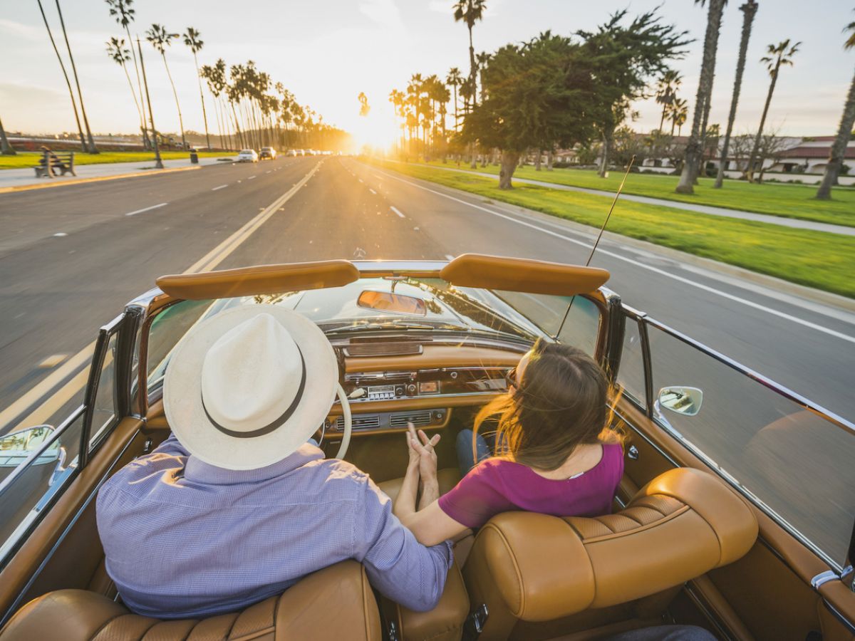 A couple drives a convertible on a scenic road lined with palm trees at sunset, holding hands and enjoying the view ahead.