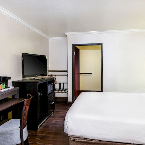 The image shows a modern hotel room with a bed, desk, chair, flat-screen TV, and a wall mirror. The room has a simple, clean, and minimalist design.