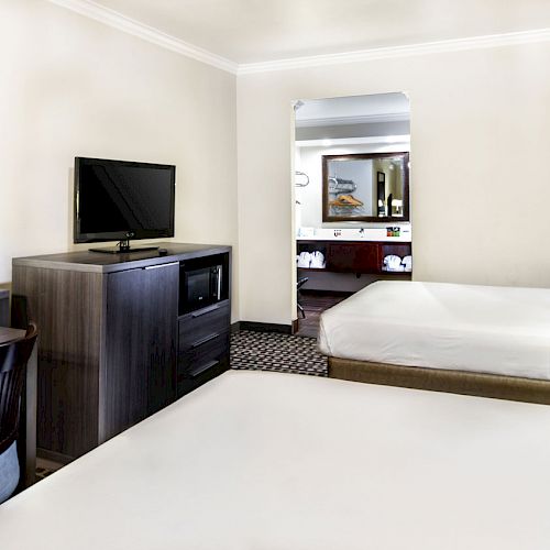 The image shows a hotel room with two beds, a TV on a dresser, a desk with a chair, and an open doorway leading to the bathroom area.