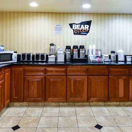 The image shows a breakfast station with wooden cabinets, a microwave, coffee makers, juice dispensers, a cereal dispenser, and other breakfast items.