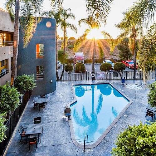 The image shows a serene outdoor pool area surrounded by lush palm trees, a building with yellow accents, and outdoor furniture as the sun sets in the background.