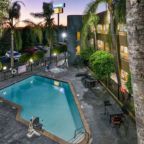 An outdoor pool area is surrounded by a building and palm trees, with lounge chairs and tables around the pool, and cars parked in the background.