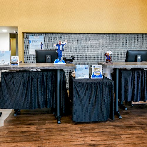 The image depicts a reception desk area with two monitors, brochures, plants, and a small figurine on the countertop, set in a well-lit room.