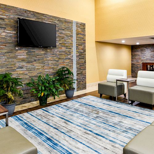 A modern lounge area with white seating, a TV on a brick wall, potted plants, a patterned rug, and high tables and stools in the back.