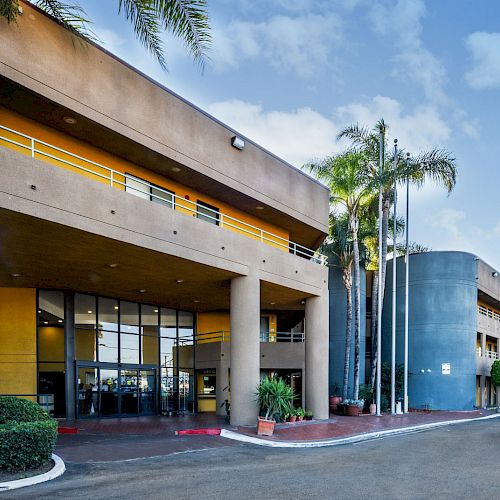 The image shows the exterior of a modern building with large pillars, an entrance, palm trees, and flags, under a partly cloudy sky.