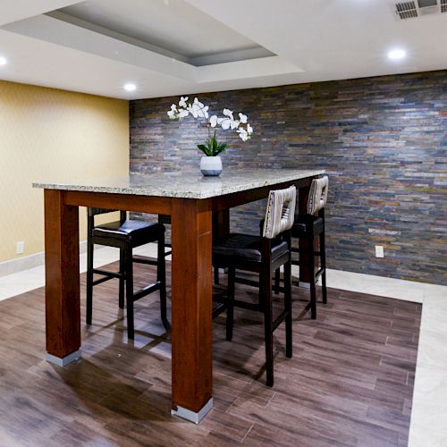 A modern room with a high table, four chairs, a small potted plant in the center, wood-paneled floor, stone accent wall, and recessed lighting.