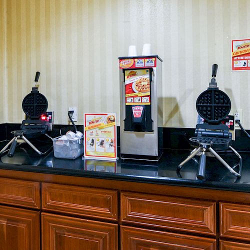 The image shows a kitchen counter with two waffle makers, a juice dispenser, and instructional signs on the wall.