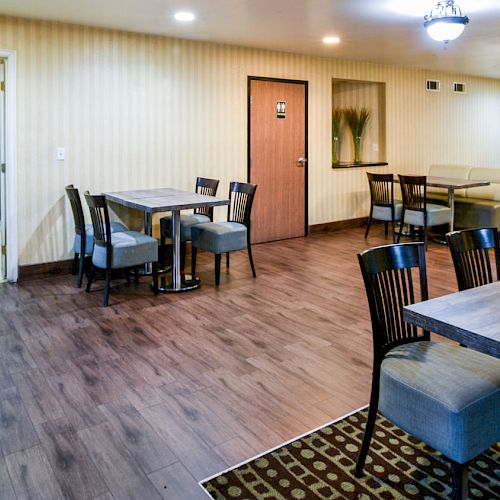 A clean dining area with several tables and chairs, adjacent to a kitchenette visible through an open doorway, featuring wooden flooring throughout.