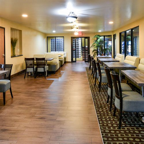 The image depicts a spacious dining area with modern wooden tables, cushioned chairs, and a mix of booth seating along the right wall and center.