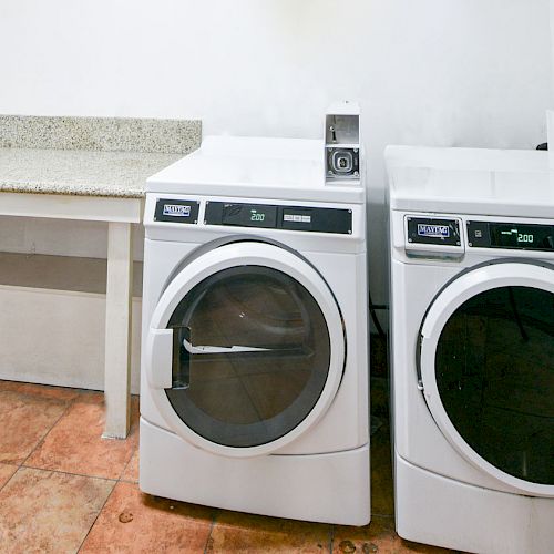 The image shows a laundry area with two white front-loading washing machines next to a countertop, with tile flooring and a white wall.