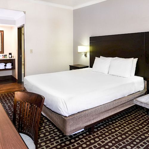 A hotel room with a bed, TV, desk, chair, and nightstands. There's an open bathroom in the background with a sink and mirror visible.