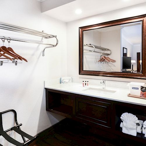 A hotel room vanity area with a large mirror, sink, hanging clothes rack with wooden hangers, luggage rack, toiletries, and coffee supplies.