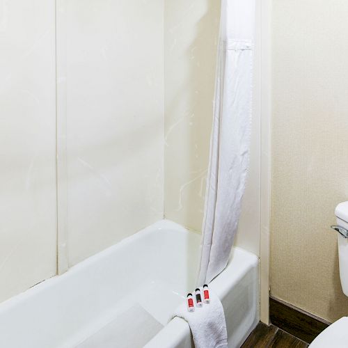 A bathroom with a white bathtub, shower curtain, and a toilet. Amenities include a towel and toiletries placed on the tub's edge.