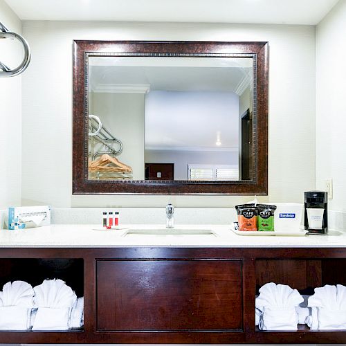 A hotel bathroom vanity with a mirror, sink, towels, toiletries, coffee maker, and cups on the countertop, with towels stored below.