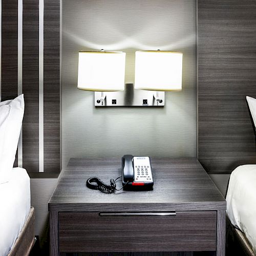 The image shows a hotel room with two beds, a nightstand in between them, a telephone on the nightstand, and two wall-mounted lamps.