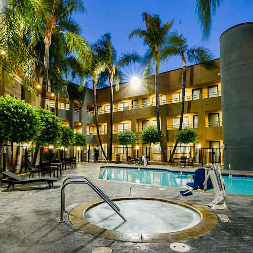 A well-lit courtyard featuring a hot tub, swimming pool, surrounded by palm trees, lounge chairs, and a multi-story building in the background ending the sentence.