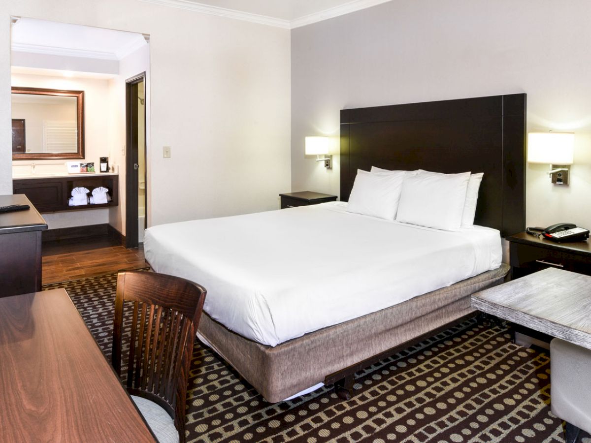 This image shows a tidy hotel room with a king-sized bed, a flat-screen TV, a desk, a chair, a loveseat, and a bathroom vanity visible in the background.