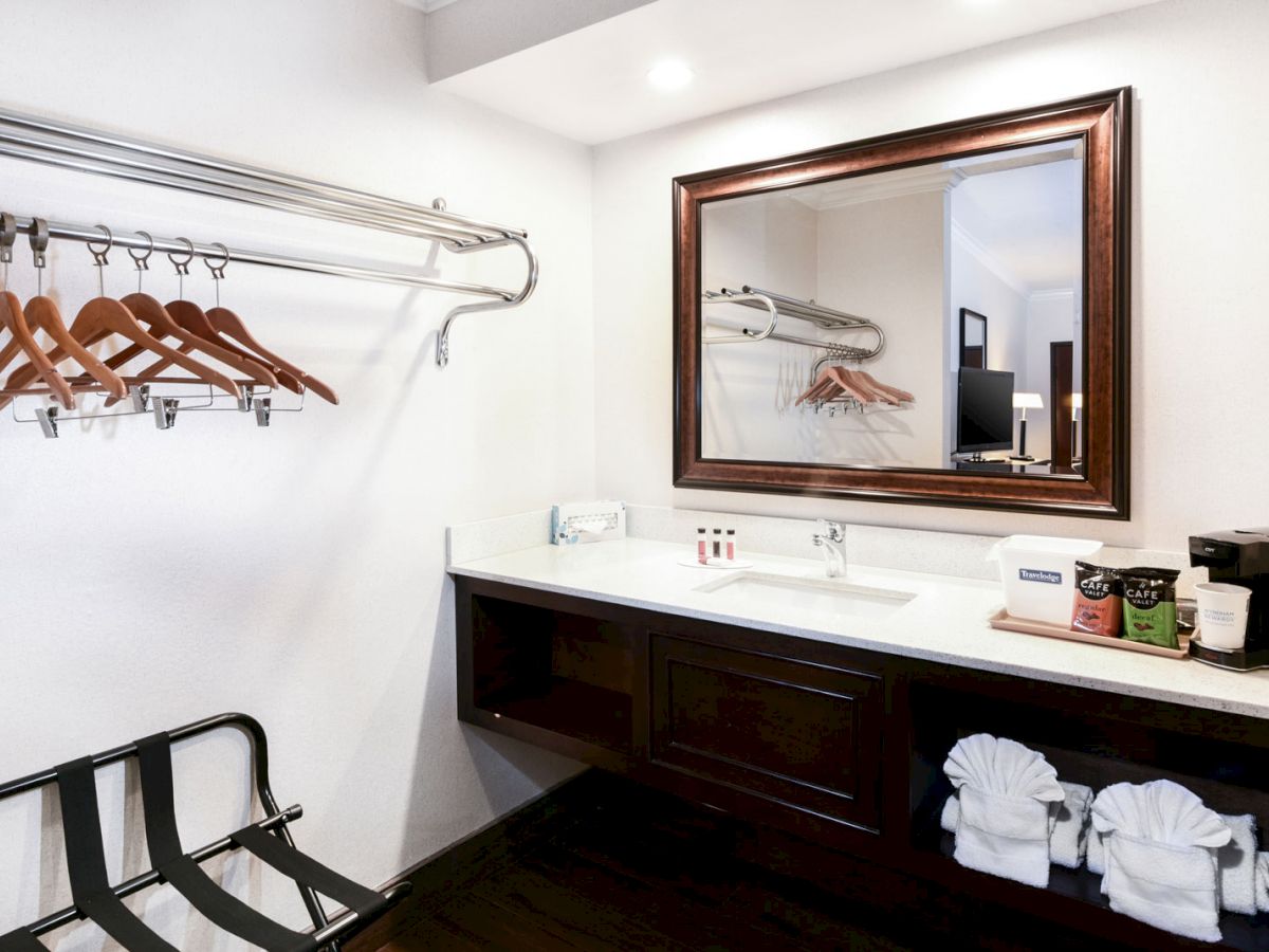 A hotel room vanity area with a large mirror, hanging rack with wooden hangers, luggage rack, sink, and various toiletries beside a coffee maker.