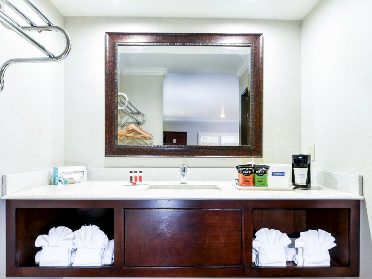 A hotel vanity counter with a mirror, toiletries, coffee maker, towels, and an overhead rack with hangers all neatly arranged in a bathroom.