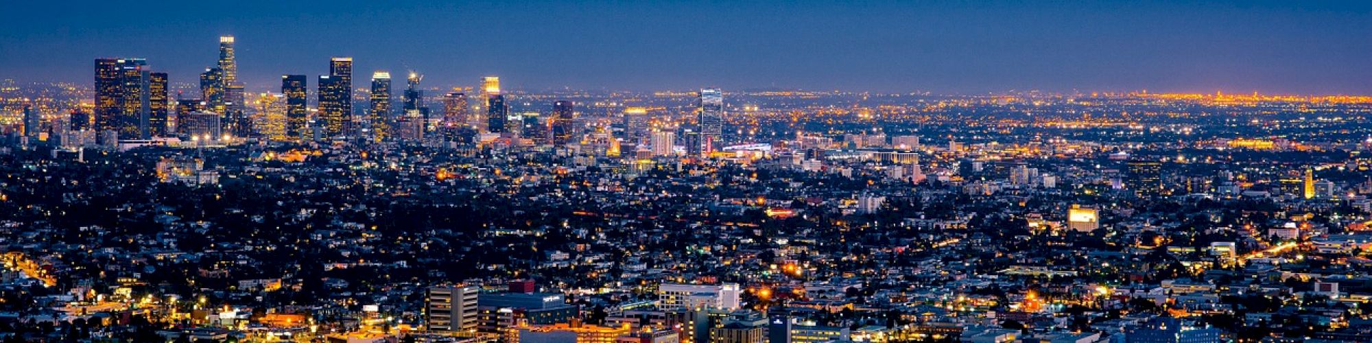 Aerial view of a cityscape at dusk, with illuminated buildings and streets stretching out to the horizon under a dark blue sky.