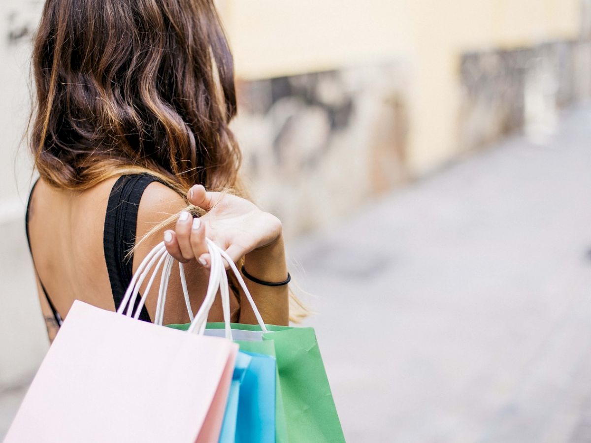 A person with long hair is walking down a street, holding several colorful shopping bags with one hand.