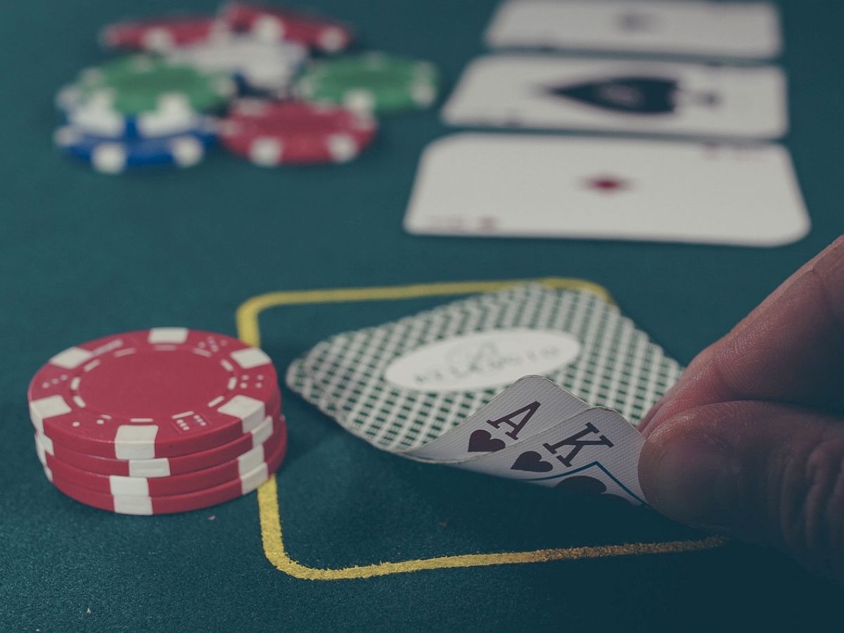 A person lifts the corner of two playing cards (Ace and King of hearts) next to a stack of poker chips on a green felt table during a poker game.