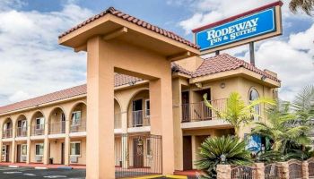 A two-story Rodeway Inn & Suites building with a prominent sign, arched windows, and a terracotta tiled roof, surrounded by palm trees and clear sky.