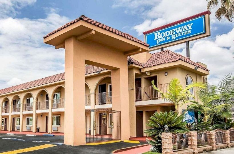 A two-story Rodeway Inn & Suites building with a prominent sign, arched windows, and a terracotta tiled roof, surrounded by palm trees and clear sky.
