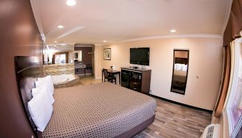 A spacious hotel room featuring a large bed, a wall-mounted TV, a small desk, a mirror, and ample lighting. The room has modern decor and wooden flooring.