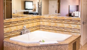 The image shows a modern bathroom with a large corner bathtub, tiled backsplash, a mounted TV on the wall, and a closed door.