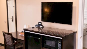 A hotel room with a TV mounted on the wall, a desk and chair, a countertop with cups and a remote, and a microwave below the counter.