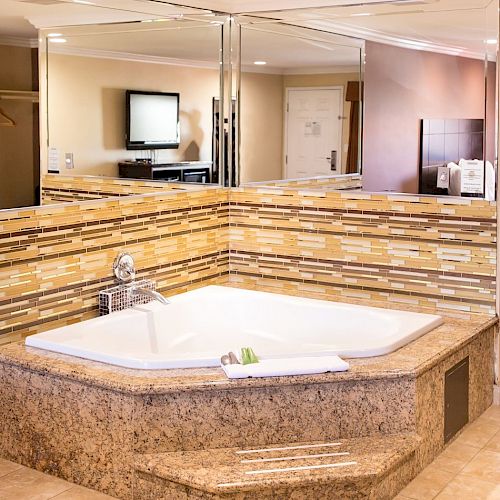The image shows a luxurious bathroom with a large, corner jetted tub surrounded by tile backsplash and mirrors, with a flat-screen TV in view always ending the sentence.
