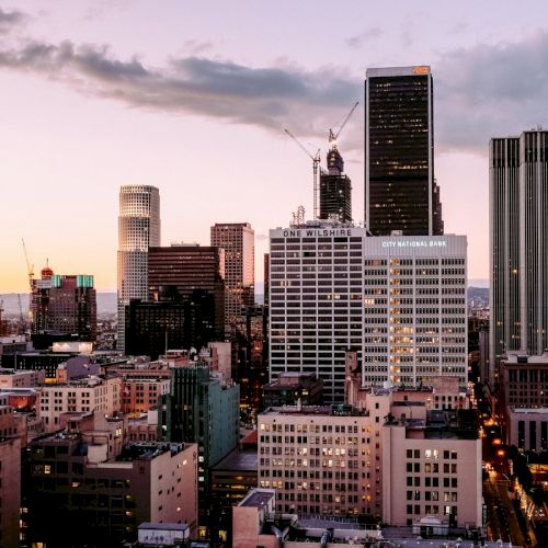 The image shows a cityscape with tall buildings and skyscrapers at sunset, with cranes indicating construction in progress under a partly cloudy sky.