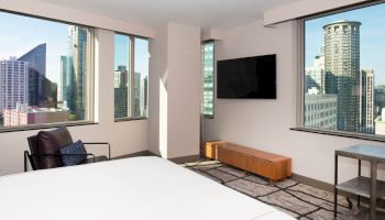 The image shows a modern hotel room with large windows, a TV, chair, and views of urban skyscrapers outside.