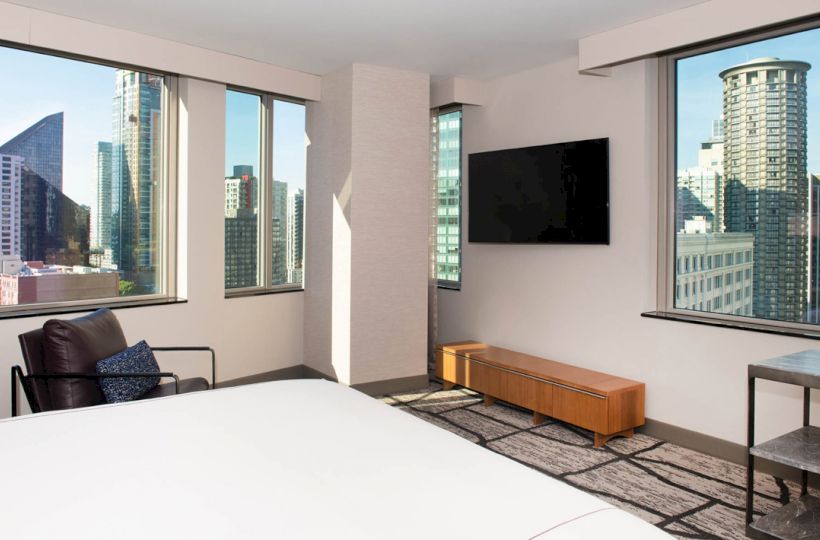The image shows a modern hotel room with large windows, a TV, chair, and views of urban skyscrapers outside.