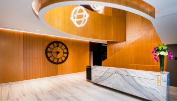 The image shows a modern, stylish lobby with a marble reception desk, wooden accents, a large wall clock, and contemporary light fixtures.