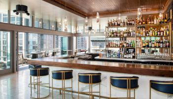 The image shows a modern bar with high-end furnishings, a well-stocked liquor shelf, barstools, large windows, and city views.
