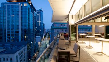 The image shows a modern cityscape viewed from a high-rise balcony with tables and chairs, overlooking a street lined with tall buildings at dusk.