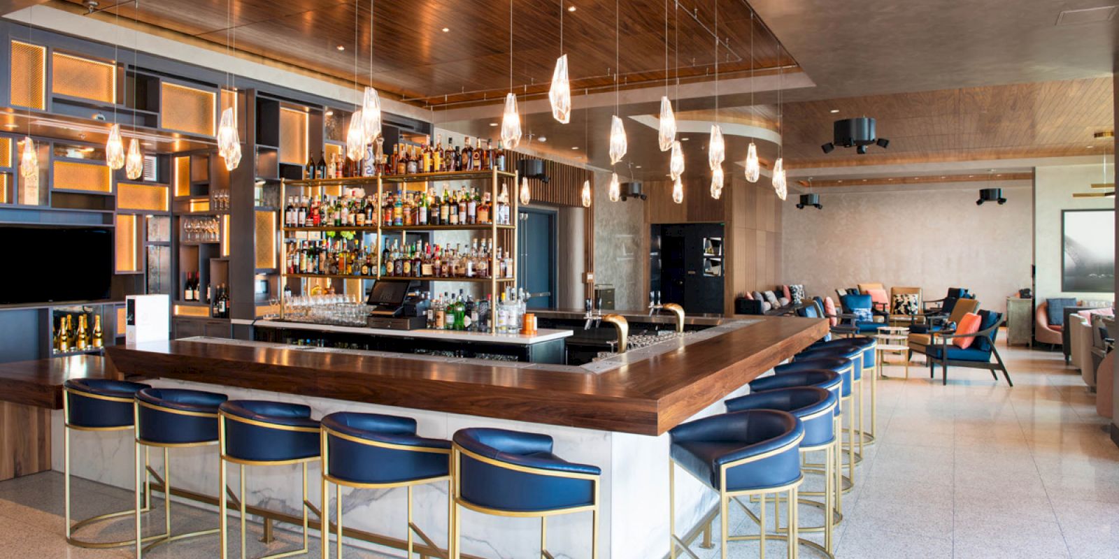 The image shows a modern bar with a wooden counter, blue barstools, and a well-stocked back shelf, featuring pendant lights and a spacious seating area.