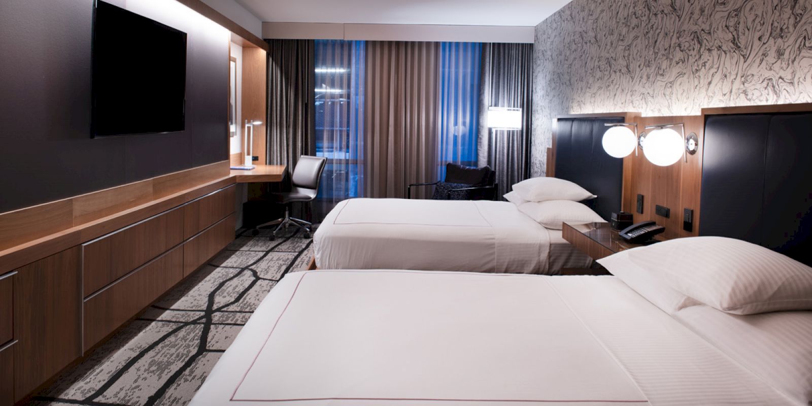 A modern hotel room with two neatly made beds, a desk with a chair, a flat-screen TV, and stylish lighting and decor.