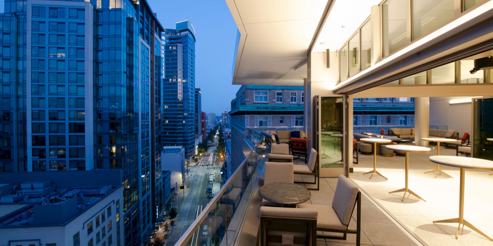 A modern, open balcony with tables and chairs overlooks a city street lined with tall buildings at dusk, featuring large windows and city lights.