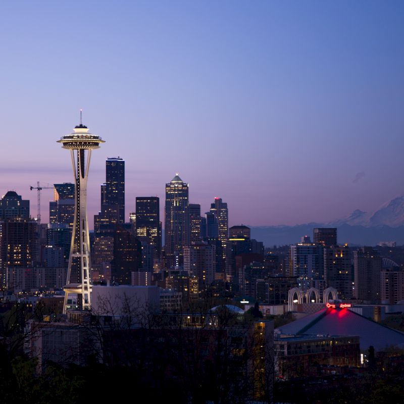 This image shows a cityscape at dusk with a prominent tower, skyscrapers, and a mountain in the background, all beneath a clear sky.