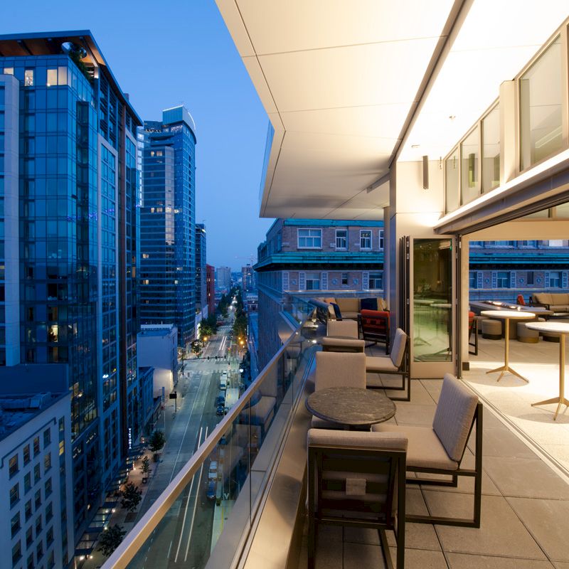 A modern, outdoor balcony scene in a city at dusk, showing tables and seating with high-rise buildings in view.