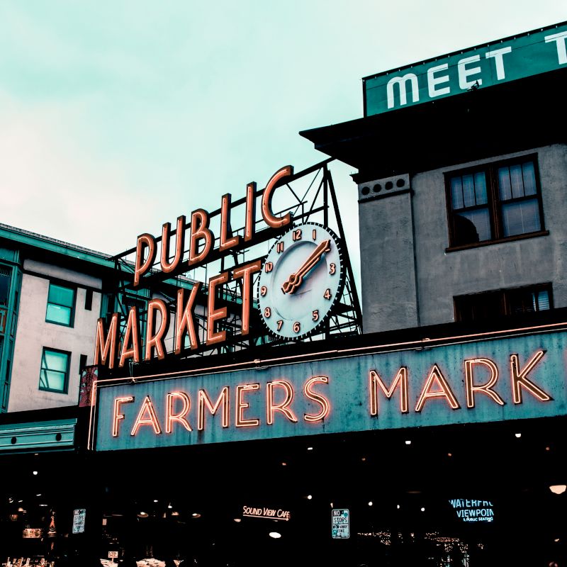 The image shows a public market with neon signs that read "Pike Place Market" and "FARMERS MARKET" against a building backdrop under an overcast sky.