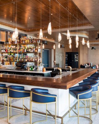 A modern bar with blue and gold stools, a central bar counter stocked with bottles, hanging lights, and a lounge area in the background.