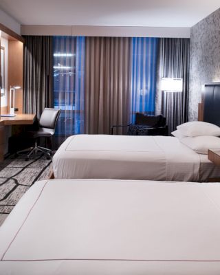 This image shows a modern hotel room with two neatly made beds, a wall-mounted TV, a desk with a chair, and contemporary decor.