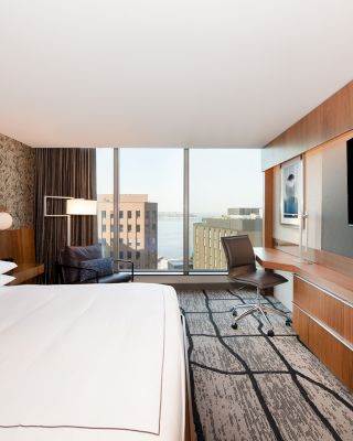 A modern hotel room with a large bed, wall-mounted TV, desk, chair, and window with a city view in the background.
