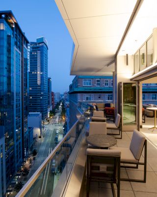The image shows a modern cityscape with tall buildings and a lit, open terrace with seating and tables, overlooking a street at dusk.